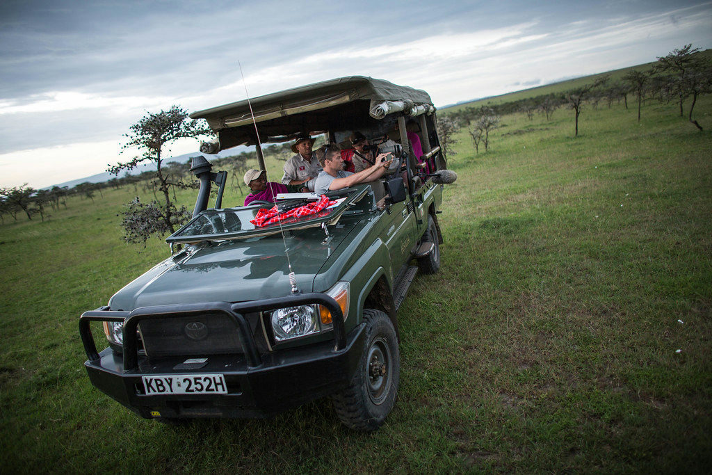 Traveler holding a smartphone beside a safari vehicle in a Kenyan savanna